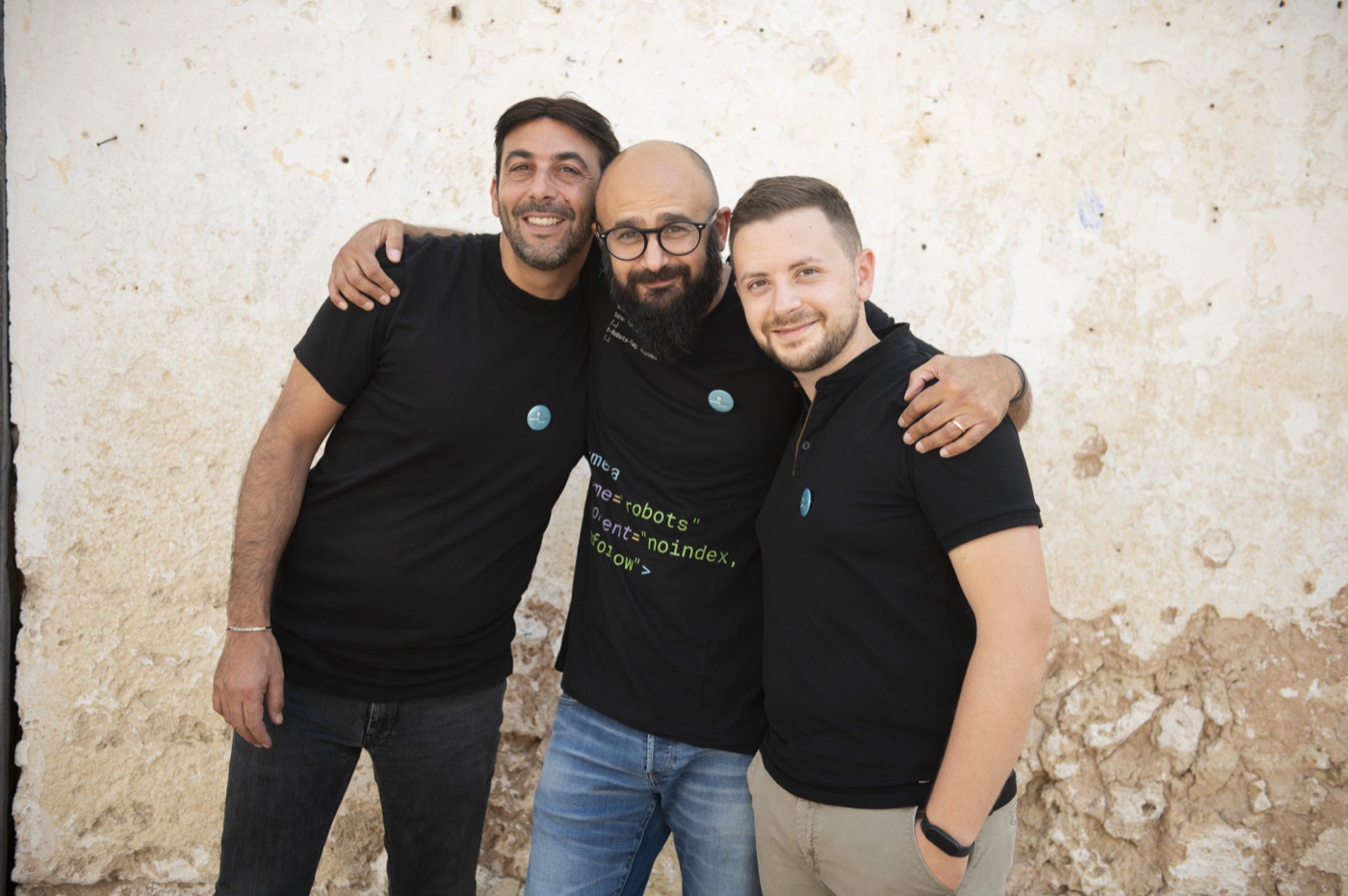 I tre soci di I/O NET abbracciati davanti a un muro di pietra, con la maglietta di Booking Designer